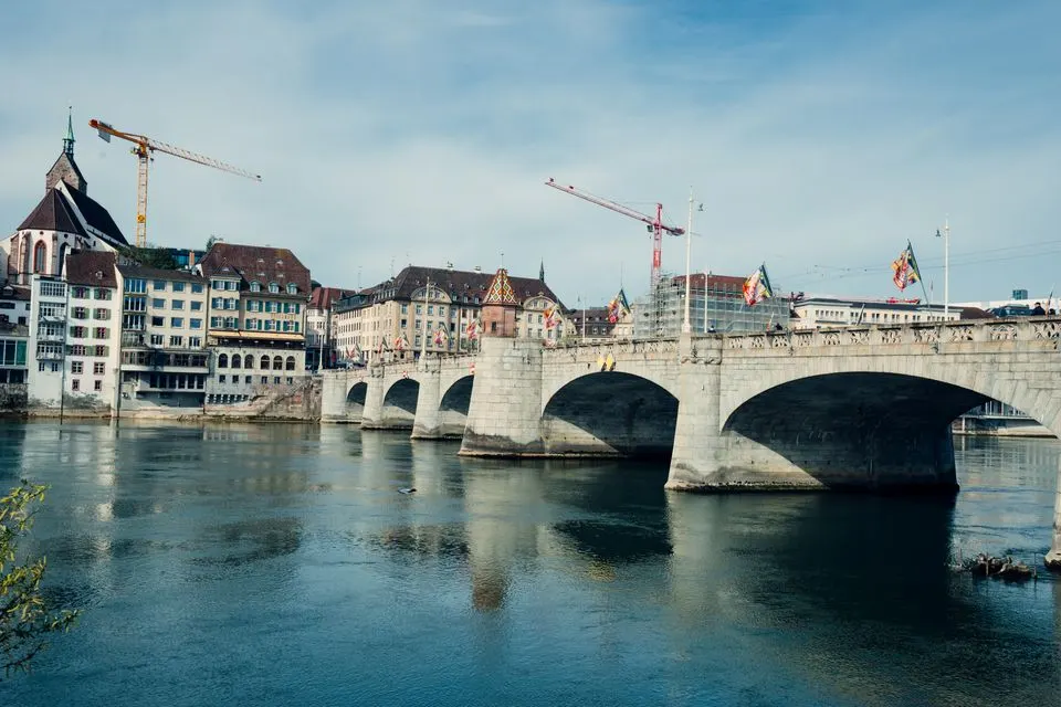 Cheval Blanc by Peter Knogl — A picturesque scene of Basel's historic bridge with city buildings and cranes by the Rhine River.