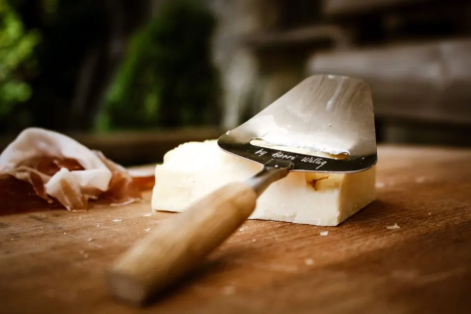 Formaggio d'alpe ticinese AOP — Close-up of a cheese slicer with cheese and prosciutto on a wooden board.