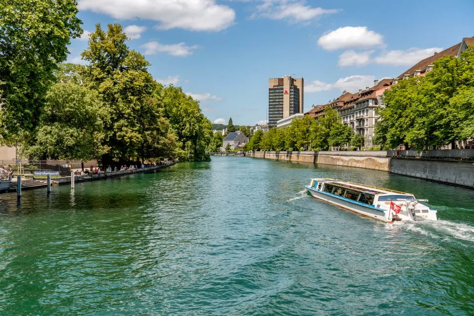 Mandarin Oriental Savoy, Zurich — A tranquil view of a river in Zurich with a boat sailing under a blue, sunny sky.
