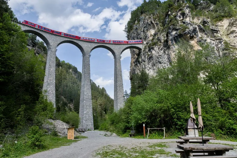 No. 4 ViaJacobi — A red train crossing the iconic Landwasser Viaduct in the Swiss Alps, surrounded by lush greenery and…