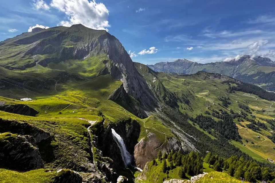 The Cambrian — Breathtaking view of Swiss Alps featuring verdant valleys, majestic peaks, and a cascading waterfall in…