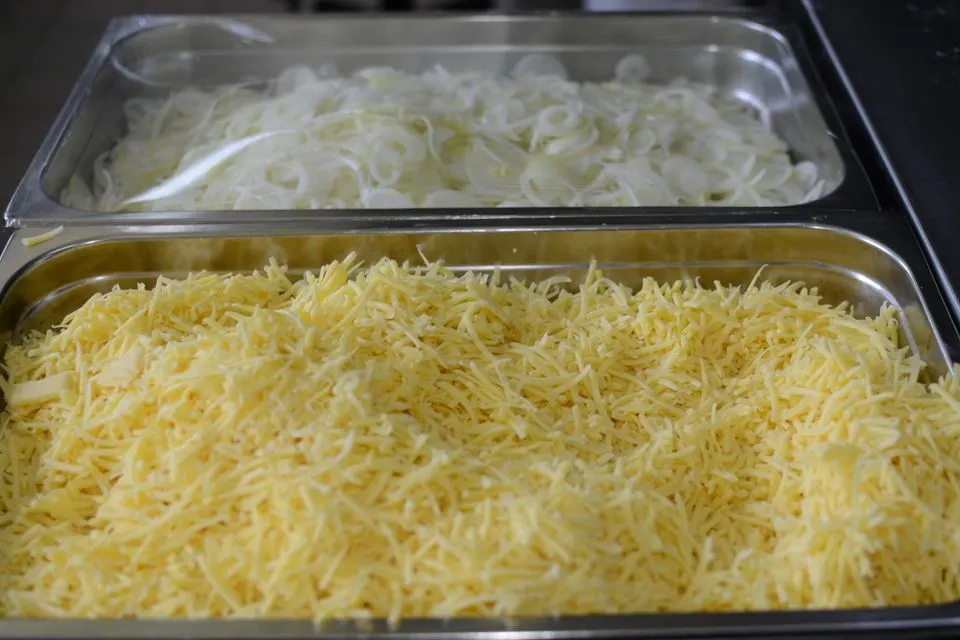 Verena — Close-up of shredded cheese and sliced onions in metal trays, perfect for cooking or catering setups.